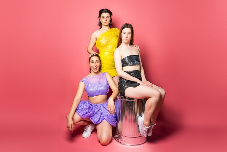 Three women posing by a shiny stool against a rosy background. 