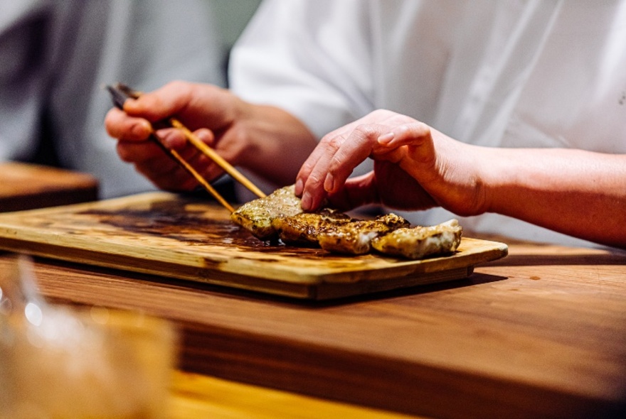 Hands holding chopsticks to lift a piece of food from a wooden board.