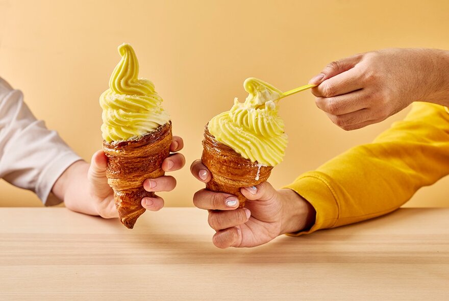 Two hands holding yellow soft serve in a pastry cone. 