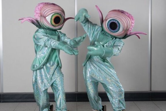 Performers in pale blue sparkly suits wearing giant eyes as heads.