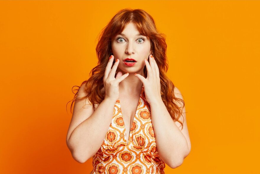 Comedian Robyn Reynolds, holding her hands to her face and looking shocked, wearing an orange patterned dress in front of an orange background.