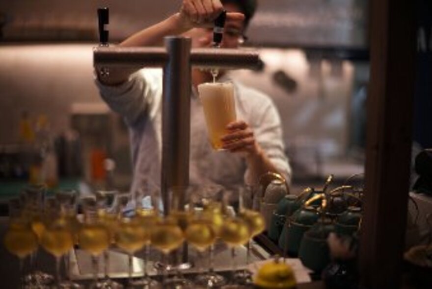 A person pouring beer from a tap into a pint glass, with more drinks in glasses in the foreground.