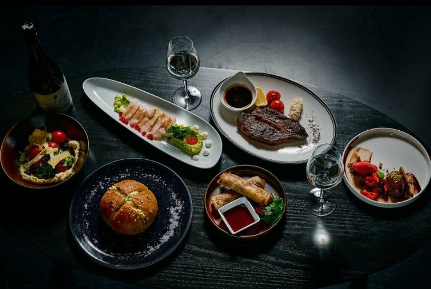 Looking down at a dark wooden tabletop with several plates of food displayed on it, and glasses of wine.