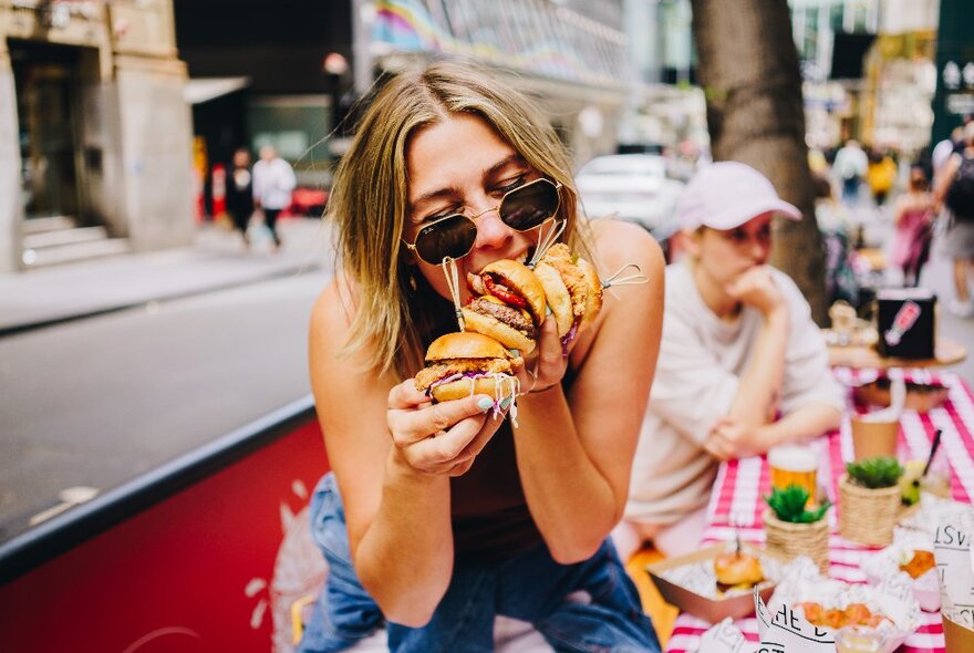 Woman wearing sunglasses trying to eat multiple burgers at an outdoor cafe.