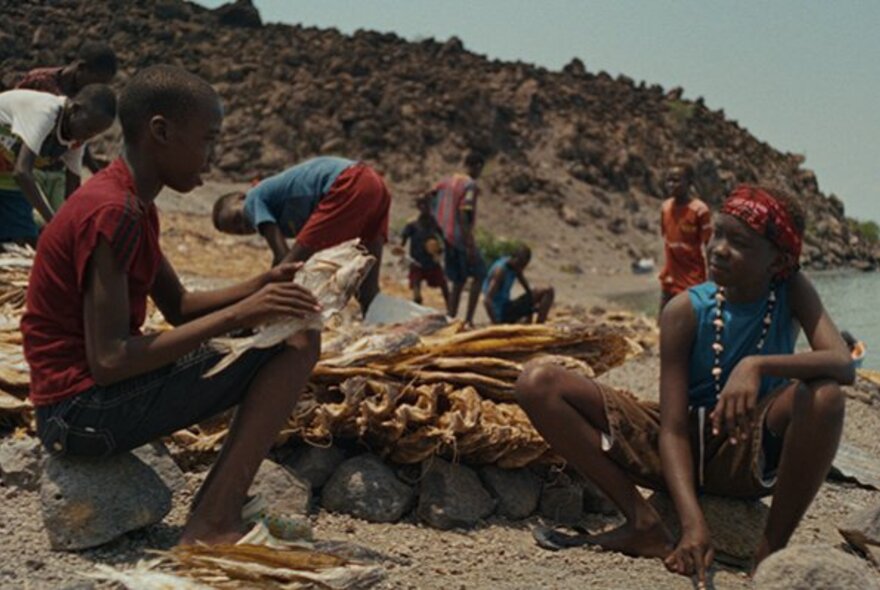 A still from an African film showing young people sitting on rocks on a beach; a collection of dried produce behind them.