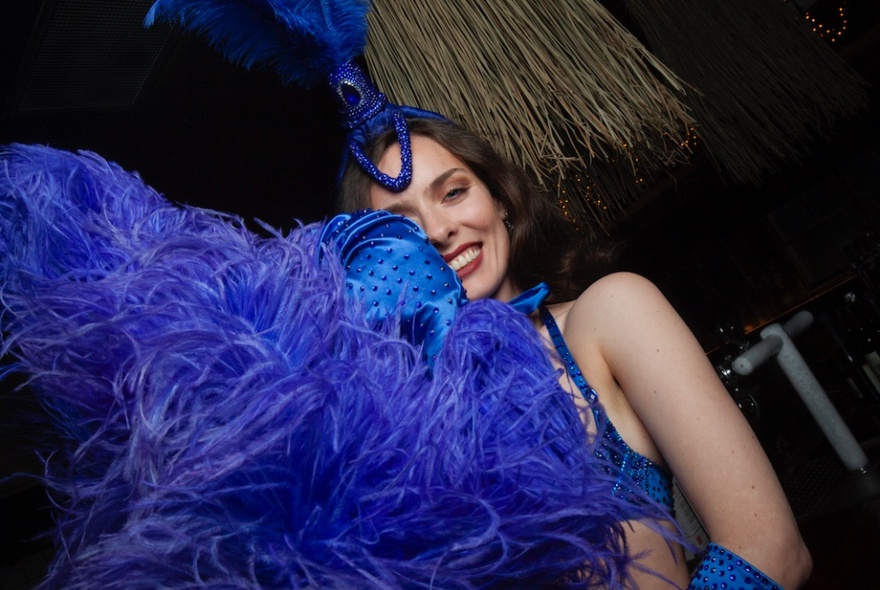 Burlesque dancer, Brook Bird, in a blue burlesque costume, smiling from behind a large blue feather boa.