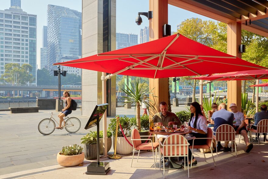 Two friends are eating and drinking alfresco style at a bar. They are sitting under a large red sun unbrella. 