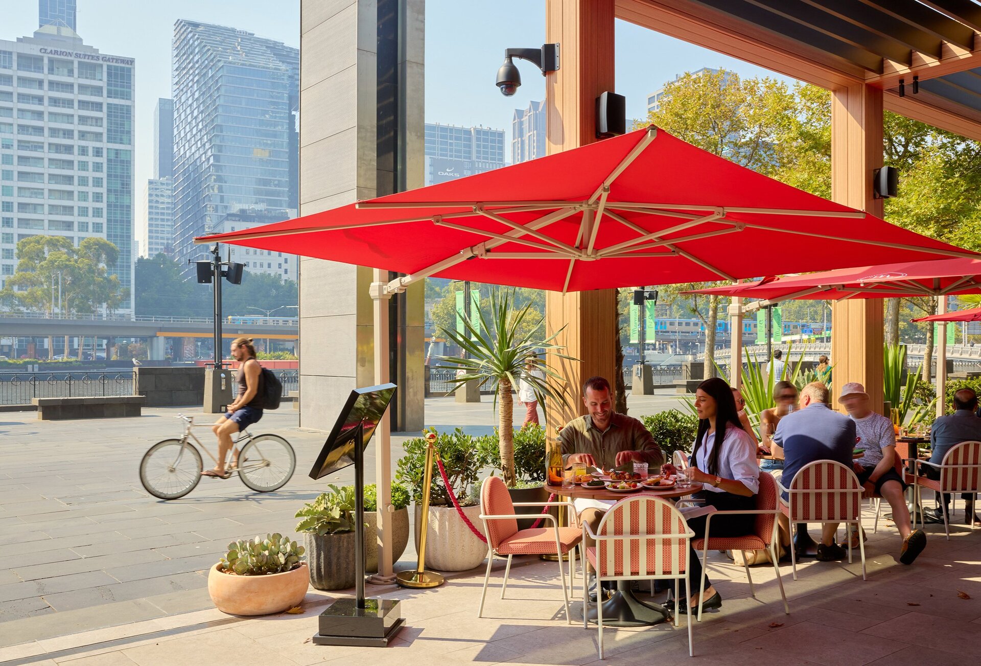 Two friends are eating and drinking alfresco style at a bar. They are sitting under a large red sun unbrella. 