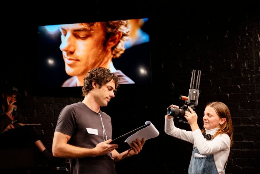 A man on stage with a clipboard being filmed by an 11-year old girl in overalls, with her photography projected onto a screen behind them.