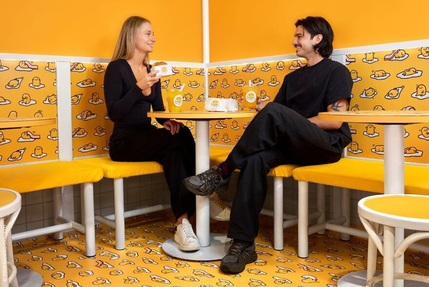 Two people seated and smiling in the yellow environs of  the Gudetama cafe.
