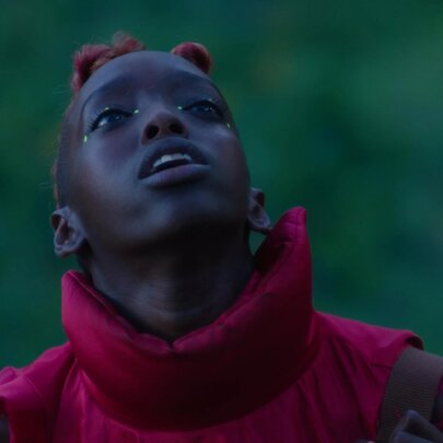 A still from the film Neptune Frost, of a young person looking up, wearing an outdoor jacket, blurred greenery behind them.