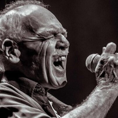 Older man singing into a microphone, his eyes closed as he reaches for a note.
