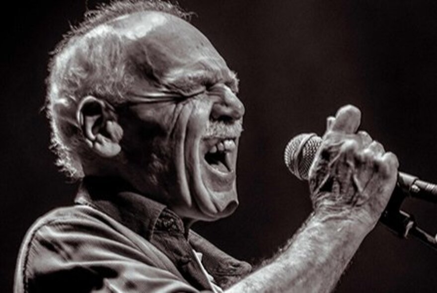Older man singing into a microphone, his eyes closed as he reaches for a note.