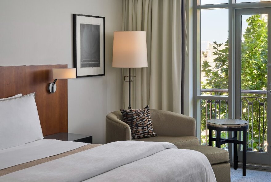 A bedroom at The Westin Melbourne, with a king size bed, a club armchair and footstool, a standing lamp, and curtains framing floor to ceiling windows.