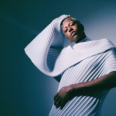 Musician Anaiis, who is looking directly at the camera while wearing a white, pleated, architectural garment with a large, voluminous collar.