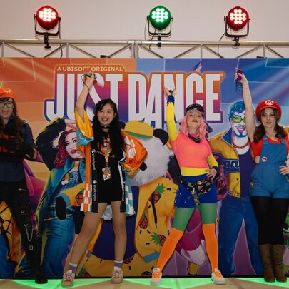 A group of people in cosplay on a stage in front of a Just Dance promotional backdrop with stage lights overhead, at an expo.