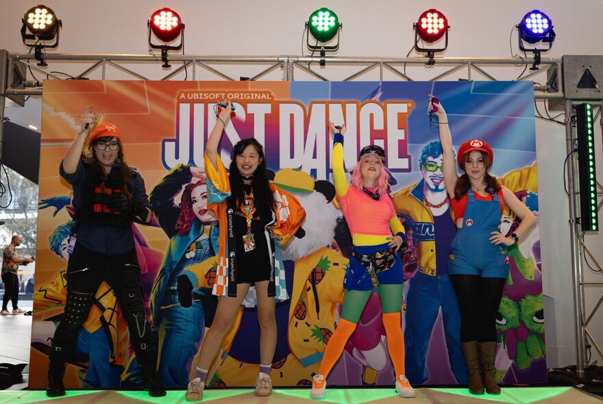 A group of people in cosplay on a stage in front of a Just Dance promotional backdrop with stage lights overhead, at an expo.