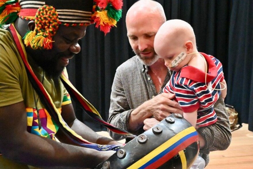 A father holding a young toddler in his arms, interacting with a man playing an African drum.