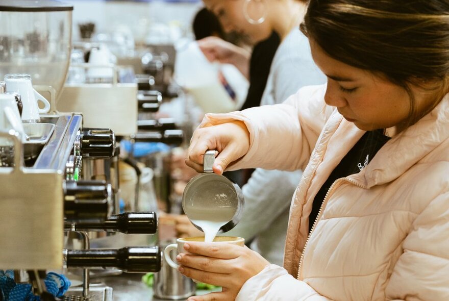 Students learning to become baristas, one woman in the foreground pouring milk into a cup. 