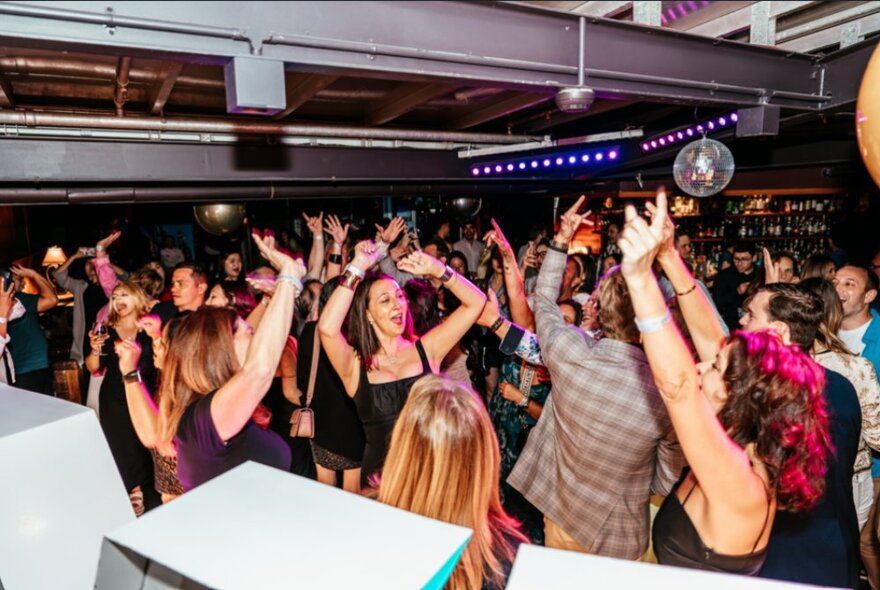 People dancing in a nightclub bar, many with their arms raised in the air.