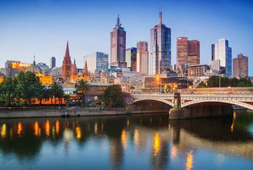 Melbourne at early dusk with lights reflected in Yarra River by Princes Bridge with buildings in the background.