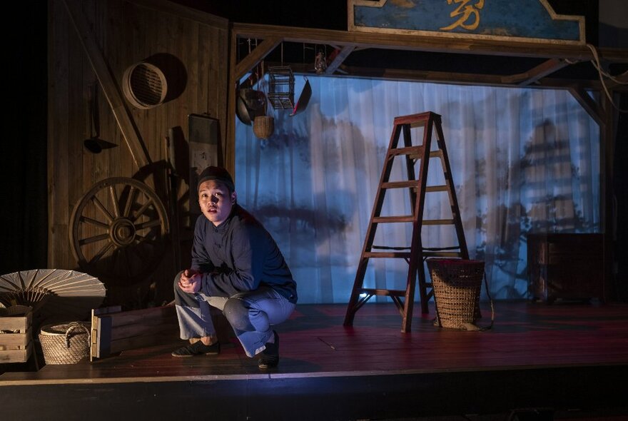 Stage set with a blue curtain, ladder and implements, with a man crouching on a stool.