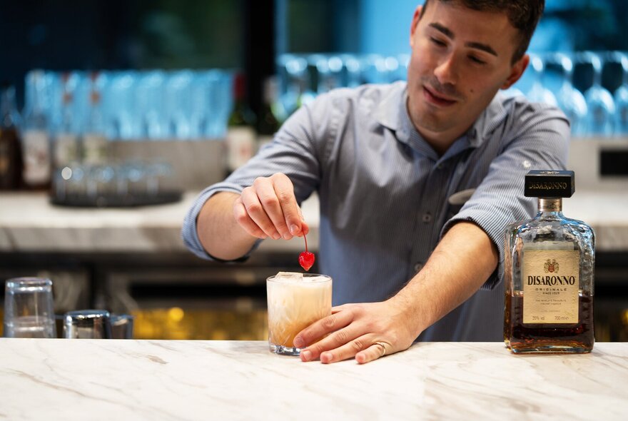 Bar tender mixing a cocktail with liqueur and maraschino cherry on a marble bar.