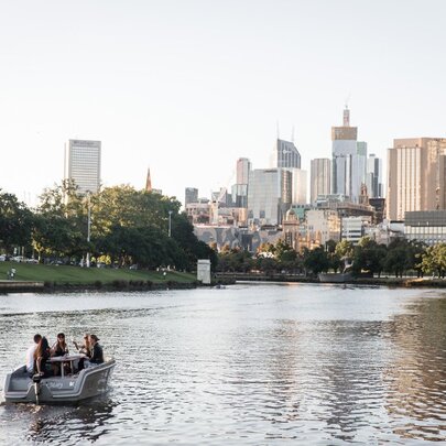 A small boat cruising along the Yarra River in the late afternoon sun, with a view of the Melbourne city skyline in the background.
