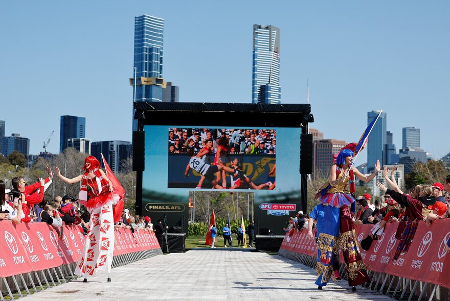 A large screen at Yarra Park as part of the Footy Fest celebrations, with roving stilt walkers interacting with the crowds that are lined up behind fence barricades.