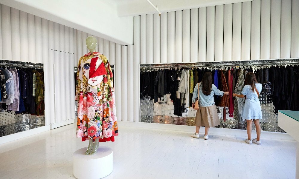 Two friends in a shop looking at clothing on racks. There is a brightly-coloured robe on a mannequin. 