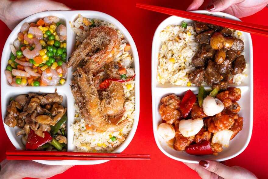 Hands holding plastic trays of Chinese meat and rice dishes with red chopsticks.