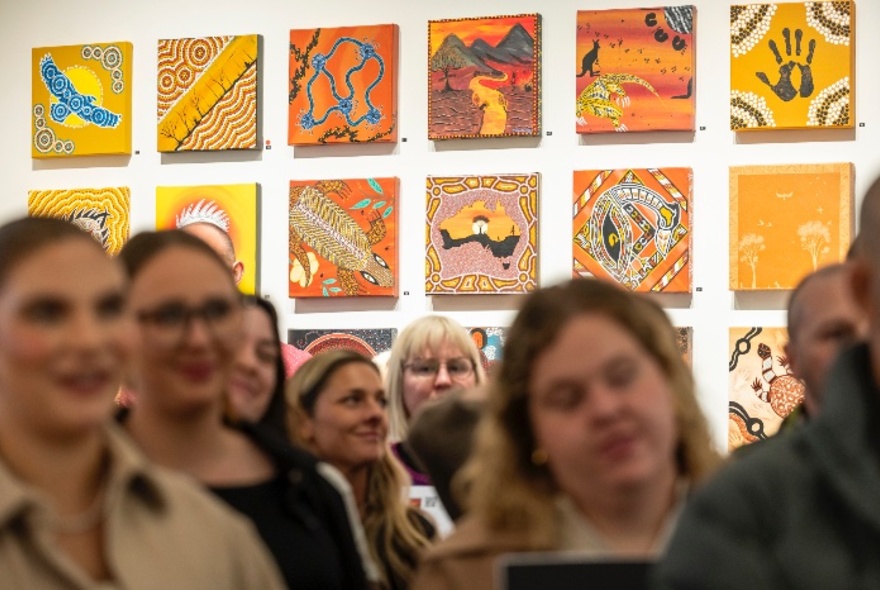 A selection of Indigenous artworks presented in a grid formation on a white wall, with an out-of-focus crowd of people in front of them.