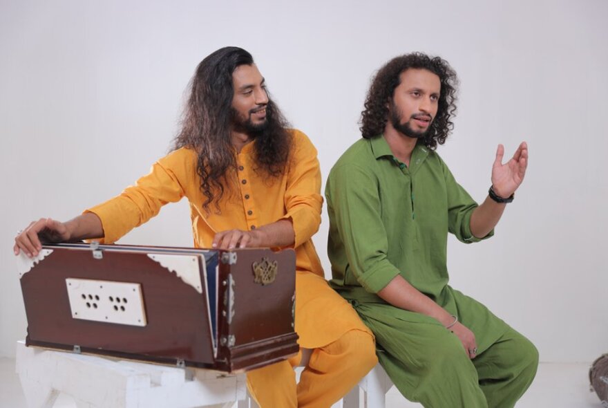 Musical group Zain Zohaib, brothers, each with long hair, one wearing all green, the other wearing all yellow and with his hands resting on a large box. 
