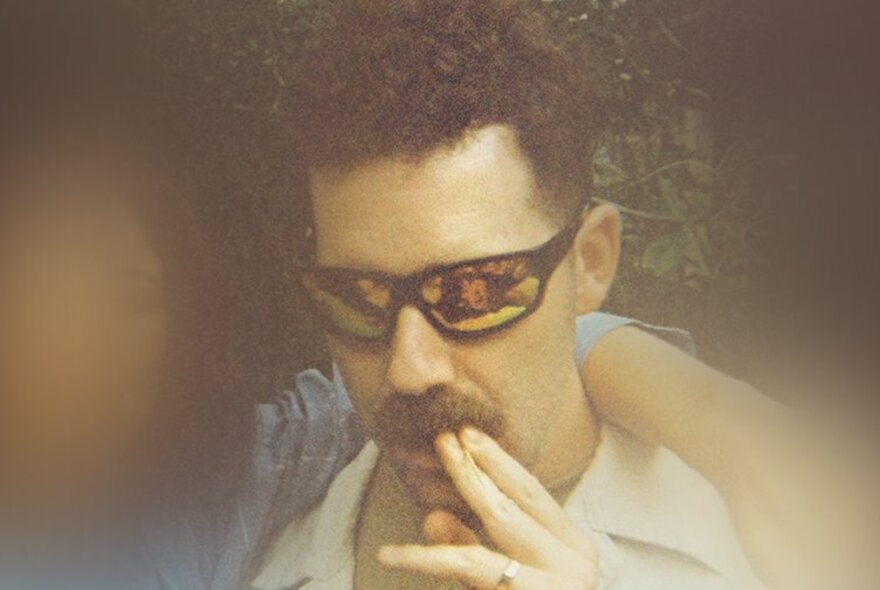 A blurry picture of a man with reflective sunglasses and a moustache, his fingers up to his lips, looking downwards. 