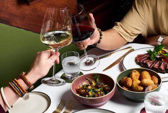 Two people clinking wine glasses and sharing salad, potatoes and steak.