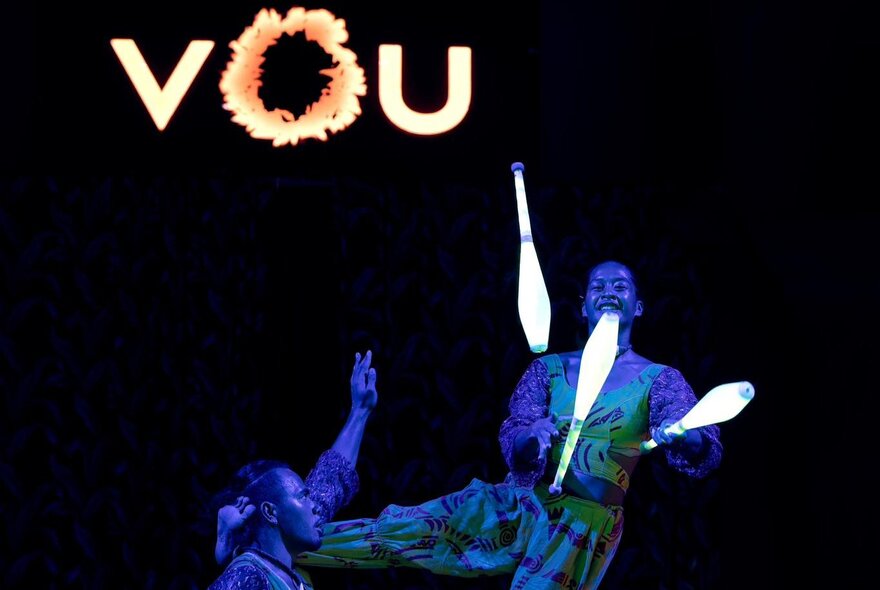 A person juggling glow-in-the-dark pins with an illuminated VOU sign in the background. 