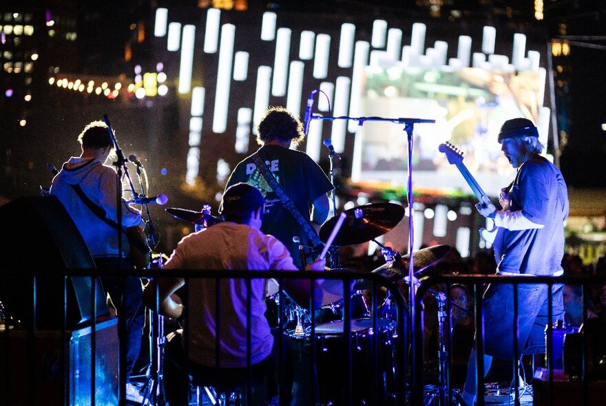 A band playing live on a stage at Fed Square, with the Fed Square digital screen in front of them; nighttime.