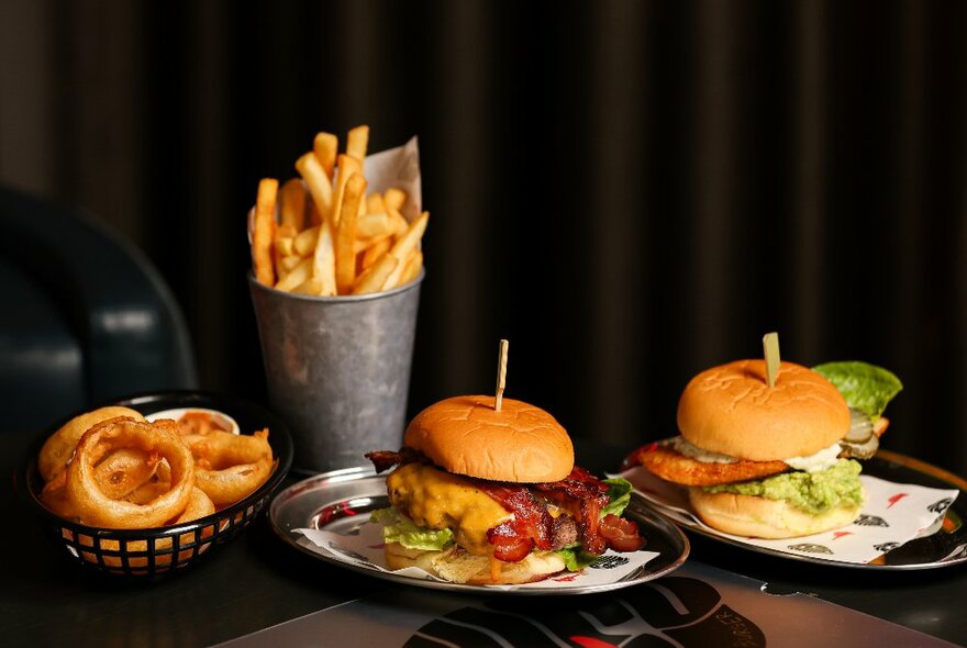 Plates of burgers, onion rings and a container of fries, on a table.