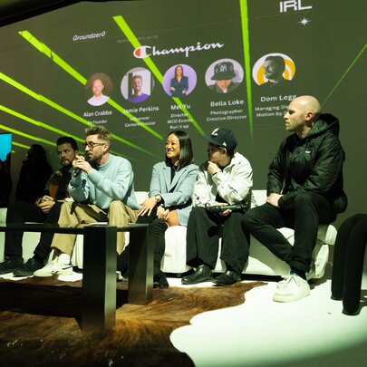 A group of speakers sit on a couch during a live panel discussion on a dimly lit stage, with a projector screen behind them showing names, circular headshots, and neon green light graphics.