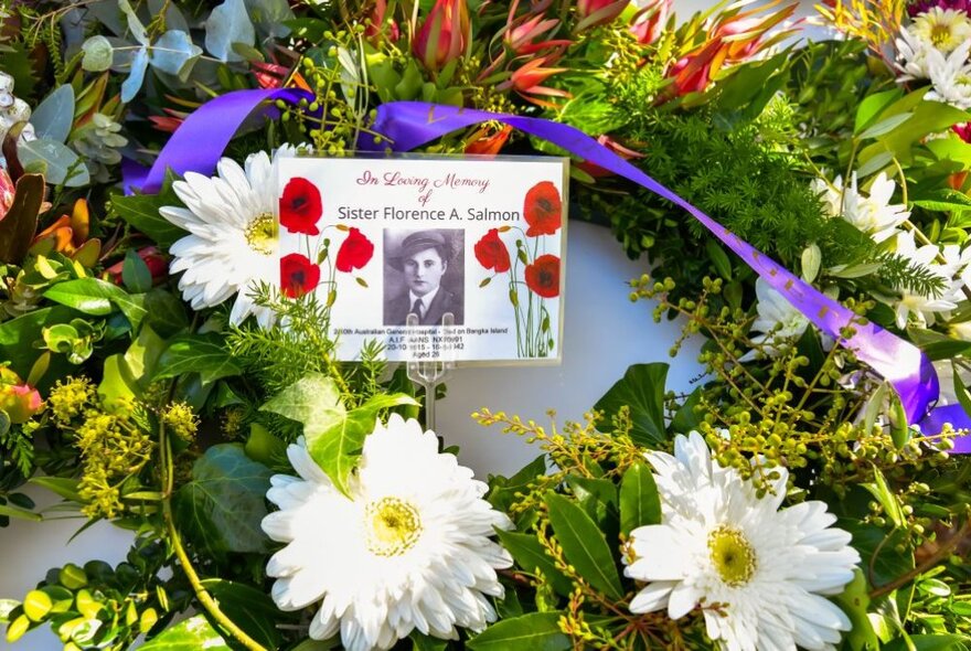 A close up of a wreath with two white Gerberas and a note with a photo of a woman and red poppies. 