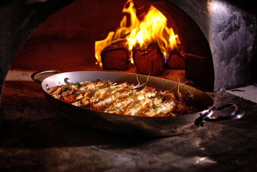 Metal platter of cooked prawns resting on a table in front of an open wood-fired oven.