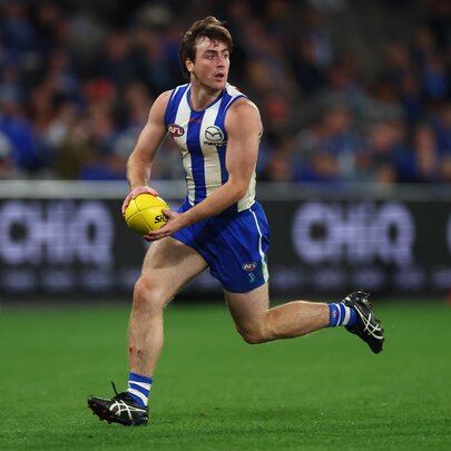 A North Melbourne AFL football player running and about to kick the ball during a match, a blurred stadium crowd in the background.