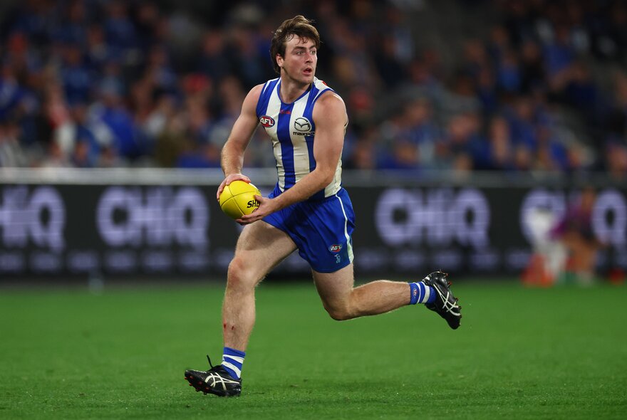 A North Melbourne AFL football player running and about to kick the ball during a match, a blurred stadium crowd in the background.