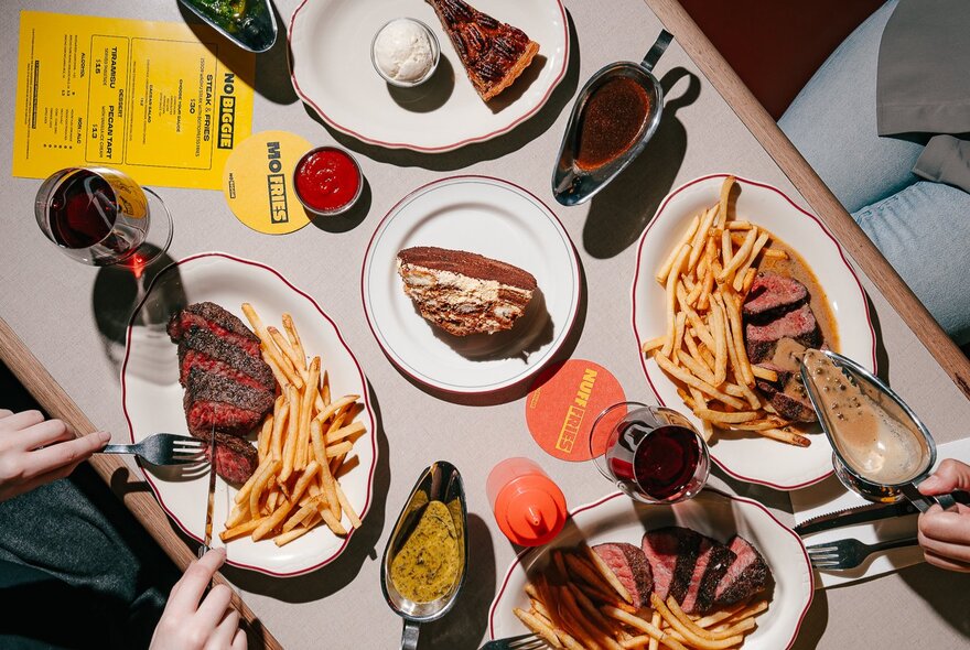 Looking down at plates of steak and fries on a restaurant table, with hands leaning in, jugs of sauce and glasses of wine. 