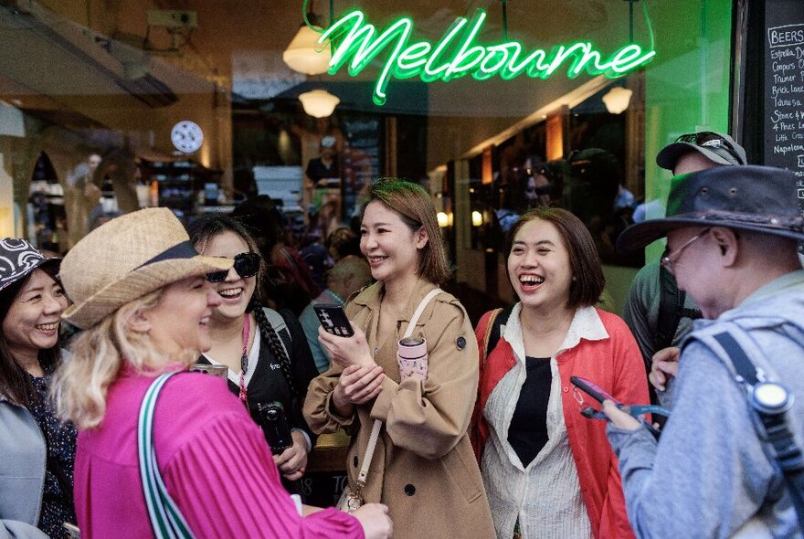 A tour group gathered in front of a green neon 'Melbourne' sign, all laughing and enjoying themselves in a huddle.