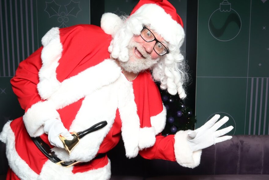 A man dressed as Santa bending over and saying hi to the camera with a wave.