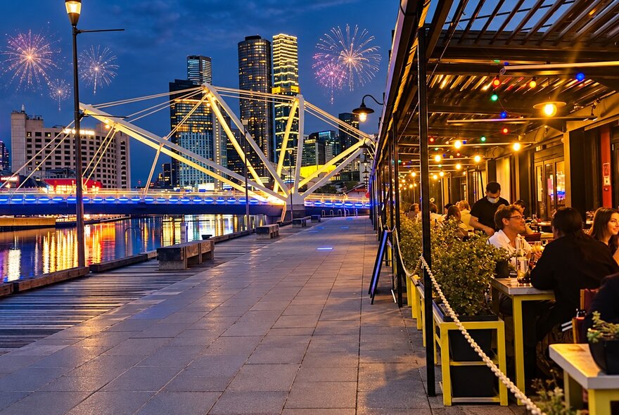 A view down the footpath at South Wharf, Melbourne, showing the outdoor tables of BangPop on the right and a lit up bridge with fireworks in the night sky.