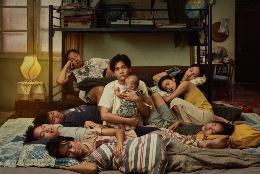 A young man seated on the floor in a small room holding a baby, surrounded by people sleeping on the floor and the couch behind him.