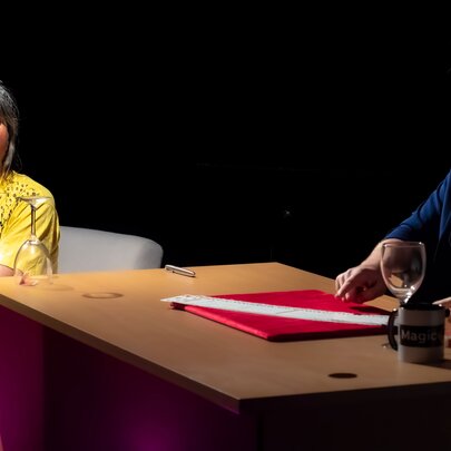 A magician seated behind a desk with an audience member watching and looking surprised.