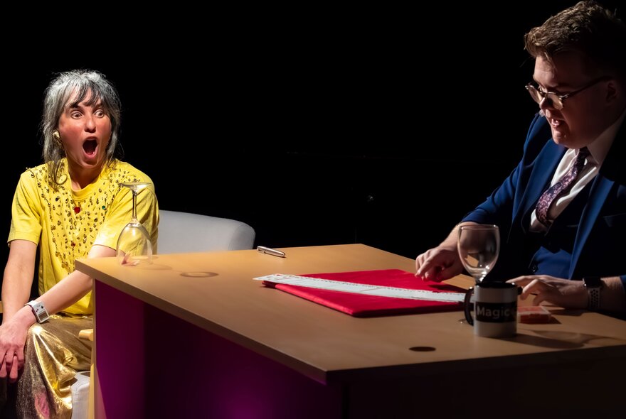 A magician seated behind a desk with an audience member watching and looking surprised.